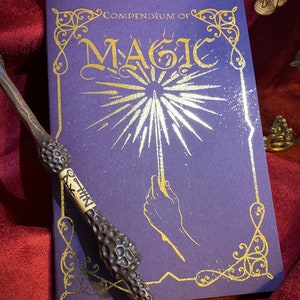 Deep purple book titled The Proficiently Practical Compendium of Magic with golden designs laid on a rich red fabric backdrop A detailed ornate wand rests diagonally atop the book. Above, a banner reads Brand new Add on. Ideal for magical enthusiasts