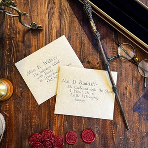 Vintage themed stationary custom parchment letters addressed to fictional characters, an intricately designed wand, round glasses, red wax seals, and an old fashioned bell on a rustic wooden table, accompanied by a twine spool and book edge.