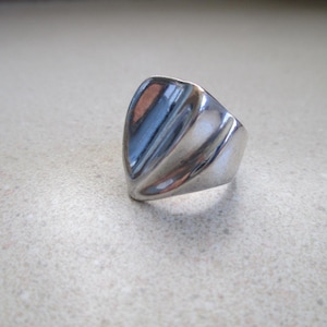 May include: A polished silver-tone ring featuring a smooth, shield-like face with three distinct vertical grooves that create a subtle, contoured texture across the surface. The band is solid and tapers toward the back of the ring.