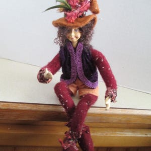 May include: A handcrafted doll with curly brown hair wearing a tall, pointed orange felt hat adorned with red leaves and feathers. The doll features a dark purple vest with decorative trim, long maroon sleeves, orange shorts, and maroon leggings.