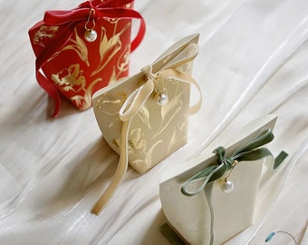 May include: Three small gift boxes with floral patterns and velvet ribbon bows. The boxes are red, beige, and white with gold accents. Each box has a small pearl charm hanging from the ribbon.