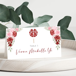 Puede incluir: Una tarjeta de mesa blanca con diseños florales rojos y rosas en las esquinas superior e inferior. La tarjeta tiene un símbolo chino rojo de doble felicidad en el centro y el texto "TABLE 1 Varna Michelle Yi" en rojo.
