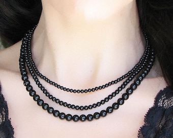 Puede incluir: Un collar de varias capas con tres hileras de cuentas negras. Las cuentas son de diferentes tamaños, creando un efecto degradado. El collar lo lleva una persona con el pelo largo y negro.