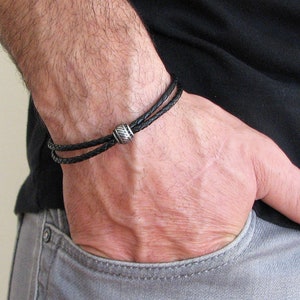 Pode incluir: Um bracelete de couro preto com um detalhe de contas prateadas. O bracelete é de dupla camada e tem um design trançado.