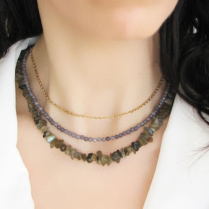 Puede incluir: Un collar de capas con una cadena de oro y dos hileras de cuentas. La hilera superior está hecha de cuentas pequeñas de color morado claro. La hilera inferior está hecha de cuentas más grandes, irregulares, de color marrón y dorado.