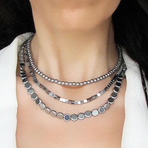 Op de afbeelding: Een gelaagde zilveren ketting met drie verschillende soorten kralen. De ketting is gemaakt van kleine ronde kralen, vierkante kralen en grotere ronde kralen.