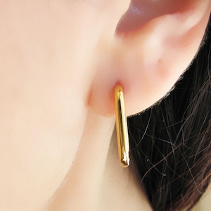 Puede incluir: Primer plano de un pendiente rectangular dorado que se lleva en la oreja. El pendiente es un rectángulo simple y alargado con bordes redondeados. El pendiente es brillante y refleja la luz.