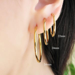 Peut inclure: Boucles d'oreilles créoles dorées de trois tailles : 22 mm, 24 mm et 34 mm. Les boucles d'oreilles sont portées sur une oreille, mettant en valeur leurs différentes tailles et une finition brillante et polie. Design simple et élégant.