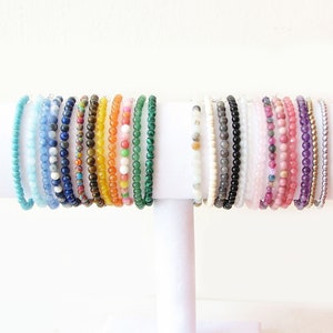 May include: A display of 20 colorful beaded bracelets on a white stand. The bracelets are made of various gemstones, including turquoise, lapis lazuli, carnelian, jade, amethyst, and rose quartz.