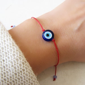 Può includere: Braccialetto di corda rossa con un ciondolo blu a forma di occhio turco. Il ciondolo ha un centro bianco e un anello blu intorno.
