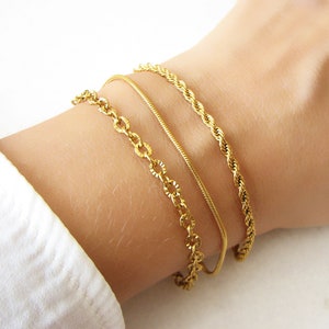 May include: Three gold bracelets stacked on a wrist. The bracelets are different styles, including a chain link, a rope chain, and a thin snake chain.