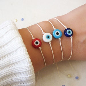 Puede incluir: Cinco pulseras de cuerda blanca con dijes de ojo turco de diferentes colores. Los dijes son rojo, blanco, azul claro y azul oscuro.