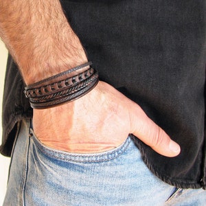 May include: A brown leather bracelet with multiple braided and woven straps. The bracelet is worn on a person's wrist.