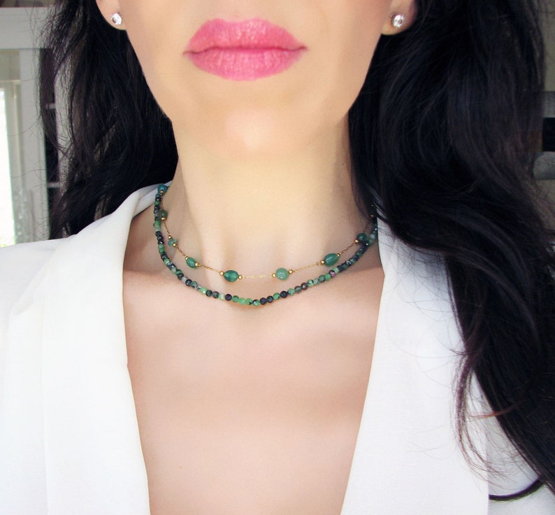 Puede incluir: Un collar de dos capas con cuentas de piedras preciosas verdes y detalles dorados. El collar lo lleva una persona con el pelo largo y oscuro.
