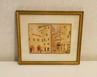 Puede incluir: Una pintura de acuarela enmarcada que representa una escena callejera en una ciudad europea. La pintura presenta edificios con puertas arqueadas y ventanas, y personas caminando por la calle. La pintura está enmarcada en un marco de color dorado.