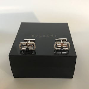 May include: A pair of silver cufflinks with a diamond-encrusted BVLGARI logo, displayed in a black box.