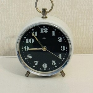 May include: A white vintage alarm clock with a black face and silver accents. The clock has a bell-shaped top and three legs. The clock face has black numbers and hands, and the words "Made in Western Germany" are printed on the bottom.