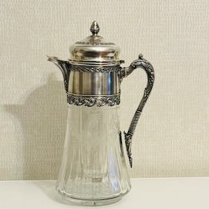 May include: A silver and glass pitcher with a decorative handle and a detailed lid. The pitcher has a clear glass body with a cut glass design and a silver top with intricate floral details.