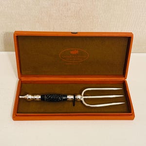 May include: A silver serving fork with a black handle, presented in an orange box with a brown interior. The box has the text "SANDRA JORDAN" on the inside. The fork has three tines and a decorative handle.