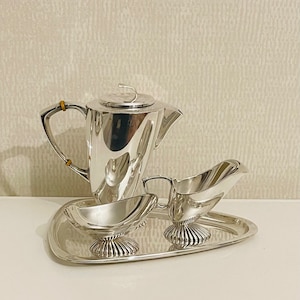 May include: A silver tea set with a teapot, creamer, and sugar bowl on a silver tray. The teapot has a curved spout and a lid with a small handle. The creamer and sugar bowl have a similar curved design.