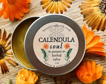 May include: A round, silver tin of "CALENDULA soul" herbal salve, with the lid slightly ajar. The label features text and illustrations of calendula flowers. The tin is surrounded by bright orange calendula flowers, and the text "organic & wildcrafted".