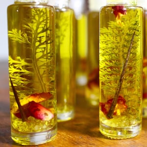 May include: Four clear glass bottles filled with a yellow liquid and dried botanicals. The bottles have a textured surface and are arranged in a row on a wooden surface.