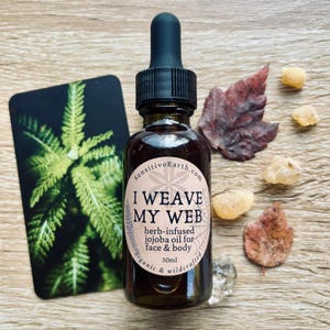 May include: A dark amber glass bottle with a black dropper, labeled "I WEAVE MY WEB" with "herb-infused jojoba oil for face & body." The bottle is 30ml. Beside it are leaves, crystals, and a card with a green plant.
