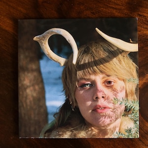 May include: A person with short blonde hair wearing a white antler headband. The person has blue and red eyes and is looking directly at the camera. The background is blurry and appears to be a forest.
