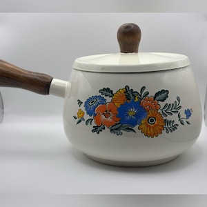 May include: A cream-colored enamel fondue pot with a matching lid, featuring a vibrant floral pattern in shades of orange, blue, and green. The pot includes a long, dark brown wooden handle and a matching wooden knob on the lid for easy handling.