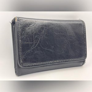 May include: A rectangular black wallet with a textured, crinkled surface finish. The wallet features a fold-over flap design with stitched edges and a slim profile, showing a consistent dark black color throughout the exterior material.