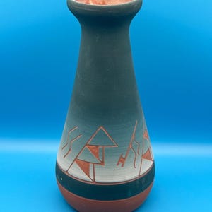 May include: A tapered ceramic vase with a matte dark green finish that transitions to a lighter grey near the base. The lower section features carved geometric patterns revealing the reddish-brown clay underneath, with a matching terracotta rim.