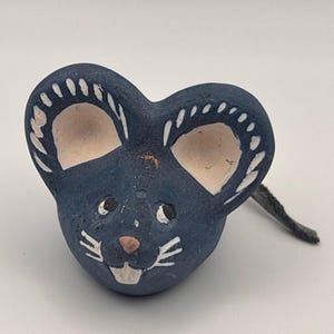 May include: A small, hand-painted blue mouse figurine with large, rounded ears featuring white decorative line details. The mouse has a light-colored snout, a small pink nose, white whiskers, and a single visible tooth. A thin, dark tail extends from the back.