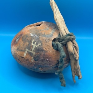 May include: A rounded, mottled brown clay vessel with a textured surface featuring a white hand-drawn symbol. A piece of light-colored driftwood is attached to the side of the vessel with a thick, dark green yarn cord wrapped around the wood.