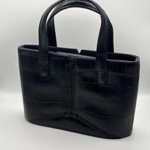 May include: A black handbag with a structured rectangular silhouette and a glossy crocodile-embossed texture. The bag features two short, fixed top handles and a central vertical seam detail on the front panel, finished with a smooth bottom edge.