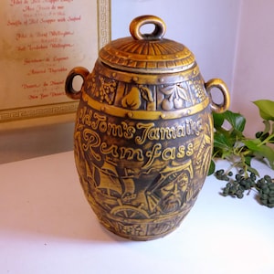 Könnte beinhalten: Ein goldfarbener Keramiktopf im Vintage-Stil mit Deckel und Griffen. Der Topf ist mit geprägten Bildern von Früchten, einem Schiff und einem Piraten verziert, mit dem Text "Old Tom's Jamaica Rumfass". Der Topf ist wahrscheinlich ein Dekorationsartikel.