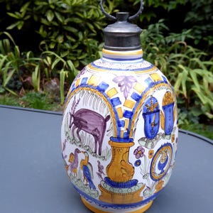 Op de afbeelding: Een keramische fles met een metalen stop en handvat, voorzien van handgeschilderde illustraties van een paars hert, vogels en bloemmotieven in geel, blauw en paars op een witte achtergrond, met een gele voet en een metalen dop.