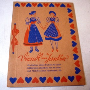 Könnte beinhalten: Eine Vintage-Broschüre mit braunem Hintergrund und einem blauen und roten Herzrand. Die Broschüre zeigt zwei Frauen in traditioneller deutscher Kleidung, mit dem Text "Dirndl und Janker" und "Den schönen echten Trachten für unsere Städterinnen abgeschaut von der Heim und Modeberaterin Indanthren-ille" auf der Vorderseite.
