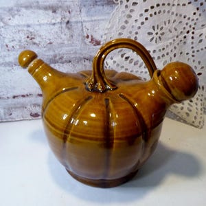 Könnte beinhalten: Ein brauner Keramikkrug mit Henkel und Ausguss. Der Krug hat eine kürbisartige Form mit vertikalen Rillen und einer abgerundeten Oberseite. Der Griff ist gewölbt und der Ausguss abgewinkelt. Der Krug ist glasiert mit einem glänzenden Finish.