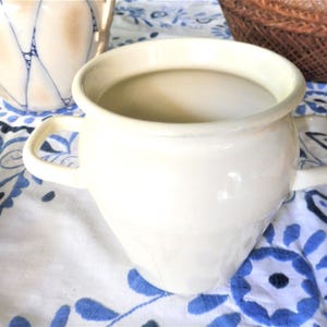 May include: A cream-colored ceramic vase with two handles. The vase has a rounded body and a wide opening. It sits on a white tablecloth with a blue floral pattern. Another vase and a woven basket are in the background.