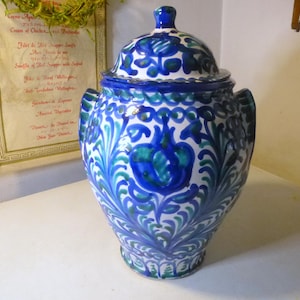 Puede incluir: Un tarro de cerámica azul y blanco con tapa, decorado con motivos florales verdes y azules. El tarro tiene un cuerpo redondeado y una boca ancha.