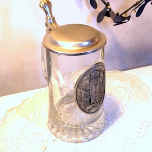 Könnte beinhalten: Klarer Bierkrug aus Glas mit einem silberfarbenen Metalldeckel und Griff. Der Krug hat ein dekoratives ovales Emblem mit einer detaillierten Illustration eines Gebäudes und dem Wort "Leobn". Der Boden hat ein strukturiertes Design.