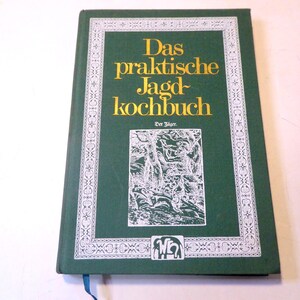 Puede incluir: Un libro de tapa dura verde oscuro con el título "Das praktische Jagdbuch" en letras doradas. Tiene un borde decorativo blanco y una ilustración de una escena de bosque. Se ve una cinta de marcador azul.