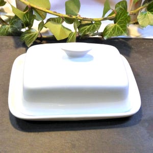 Op de afbeelding: Een witte keramische boterdoos, rechthoekig met afgeronde hoeken en een klein handvat op het deksel. De schaal staat op een bijpassende basis. Een eenvoudig, elegant ontwerp voor keuken of eetkamer. Afmetingen in cm.