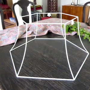 Può includere: Un telaio di paralume in metallo bianco con un design geometrico. Il telaio è composto da linee rette e angoli, creando una forma esagonale. Il telaio è appoggiato su un tavolo di legno scuro.