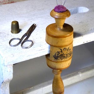 Könnte beinhalten: Ein hölzernes Nadelkissen im Klemmstil mit einem lila Kissenaufsatz, der mehrere silberne Nadeln hält. Ein kleiner Messingfingerhut und eine Vintage-Schere befinden sich neben dem Nadelkissen. Das Nadelkissen hat eine dekorative Illustration.