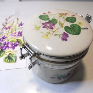 Può includere: Un barattolo di ceramica bianca con un coperchio incernierato che presenta un motivo floreale di fiori viola e gialli con foglie verdi. Il barattolo ha una chiusura in metallo argentato.