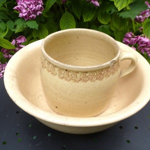 Può includere: Un set di due ciotole in ceramica vintage, una annidata nell'altra. La ciotola più grande è di colore giallo pallido e ha un bordo leggermente ondulato. La ciotola più piccola è di colore giallo pallido con una fascia decorativa marrone intorno alla parte superiore. Entrambe le ciotole hanno un aspetto rustico e fatto a mano.