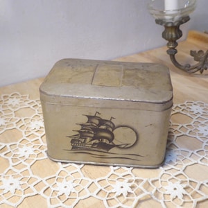 Könnte beinhalten: Eine rechteckige Metallbox im Vintage-Stil mit verblasstem Gold-Finish. Die Box zeigt eine detaillierte Illustration eines Segelschiffs mit wehenden Segeln und einer untergehenden Sonne. Die Box dient wahrscheinlich zur Aufbewahrung.