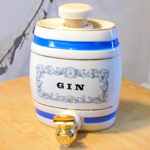 Könnte beinhalten: Weißer Gin-Fassbehälter aus Keramik mit blauen Streifen und goldenen Akzenten. Der fassförmige Behälter hat einen Zapfhahn und einen Korken. Das Wort "GIN" ist in schwarzer Schrift auf der Vorderseite zu sehen.