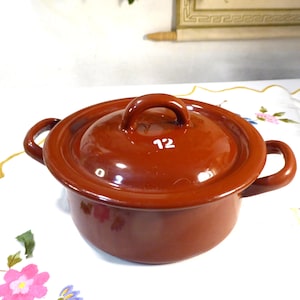 Op de afbeelding: Een kleine, ronde, bruine emaille pot met een bijpassende deksel. De deksel heeft een lusvormige handgreep en het nummer "12" in wit. De pot heeft twee zijhandgrepen. De pot staat op een tafelkleed met bloemenpatroon.